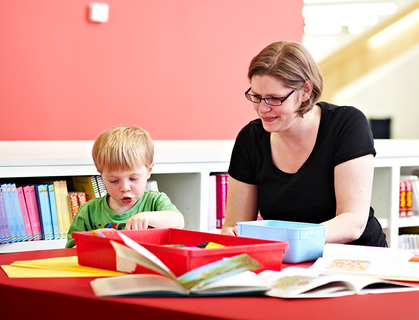 Young child and adult woman sitting at a red table with books in a bright learning space.