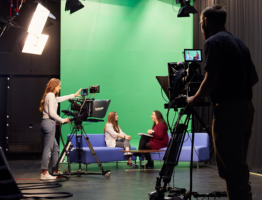 Film studio with green screen. Two students sit on sofas talking whilst another two students stand behind large cameras filming