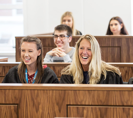 Students in black robes sat in wooden stall in the mock courtroom. They are smiling and laughing.