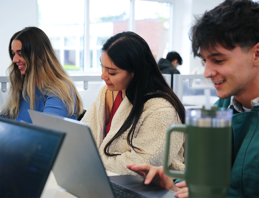 3 students sat beside each other smiling whilst working on laptops