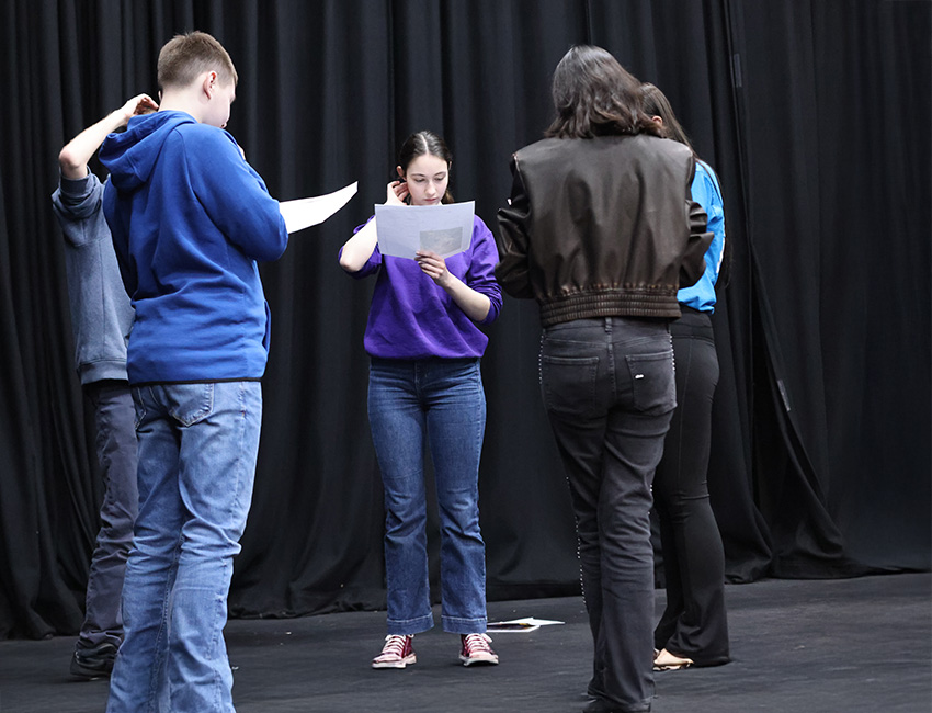 Students stood in a circle in a drama studio, reading a script.