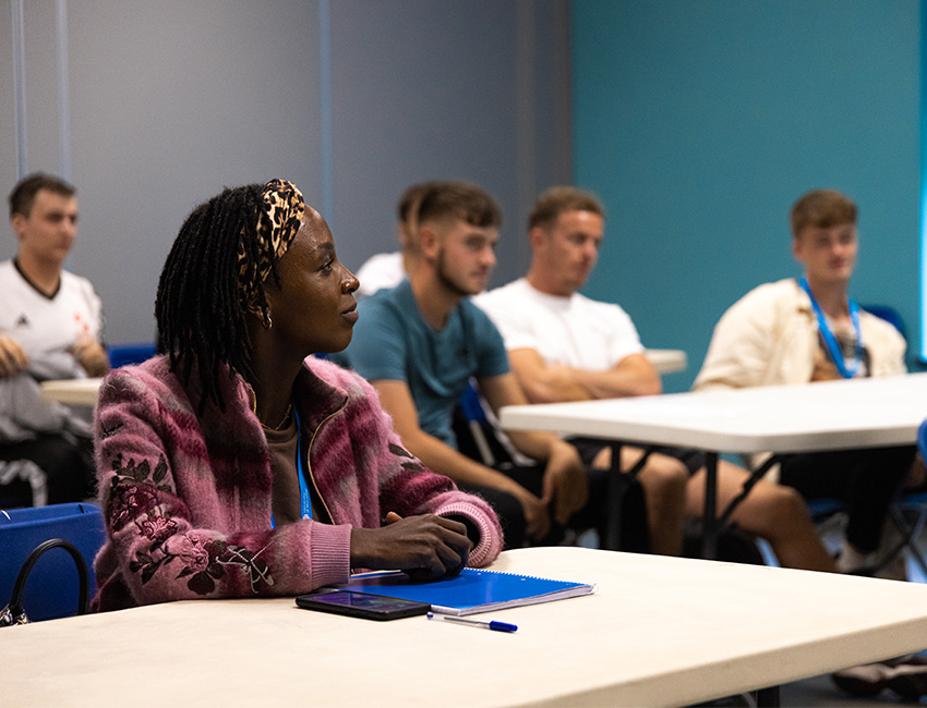 A student listening to a lecture whilst sat at a desk with a notebook and pen