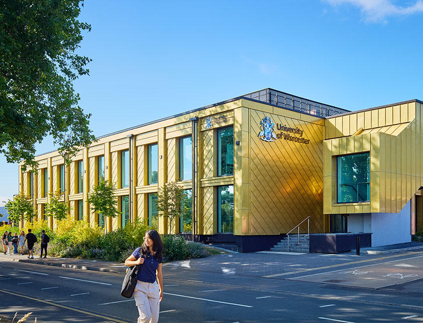 A modern gold‑clad university building with large vertical windows on a bright sunny day, with the University of Worcester logo visible. Trees and landscaped planting line the pavement, and a person walks along the street in the foreground.