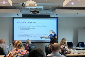 Woman giving a presentation to a people in a meeting room with presentation projected on to a screen.