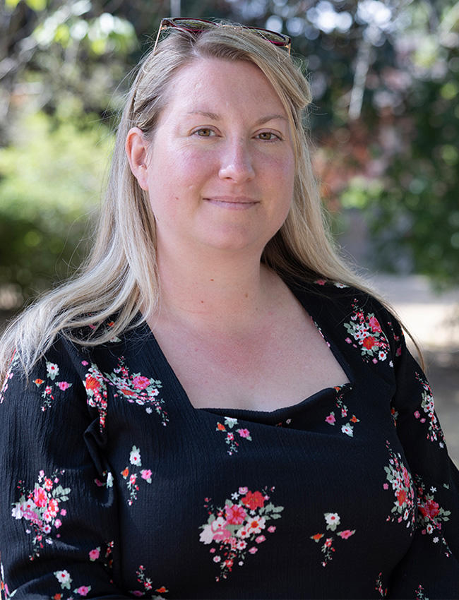 Photo of Hannah, wearing a black floral dress and stood outside with greenery behind her