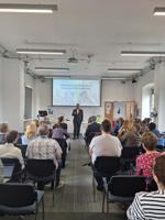 A group of conference delegates watch a speaker in front of a digital screen