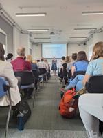 A group of conference delegates watch a speaker in front of a digital screen