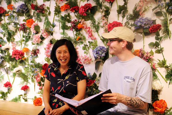 Two people sit on a bench with a folder against a floral backdrop