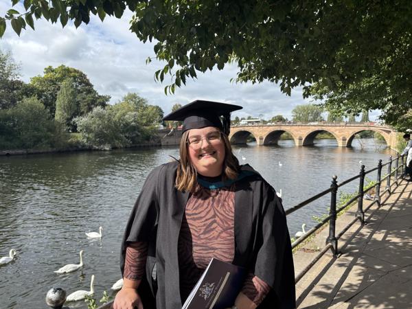 Abbie poses in her graduation cap and gown by the river in worcester