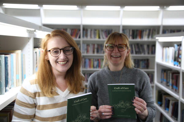Authors Charley Barnes and Claire Walker pose among books in a library with a copy of their books, smiling at the camera