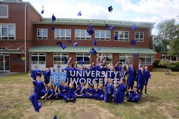 Around 30 children in purple robes throw their graduation caps in the air outside around a University of Worcester sign
