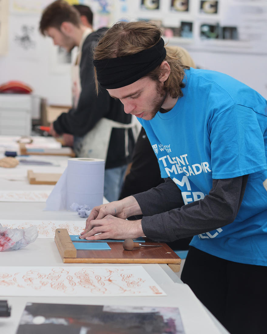 A student works on his artwork in a blue tshirt at a white desk covered in art