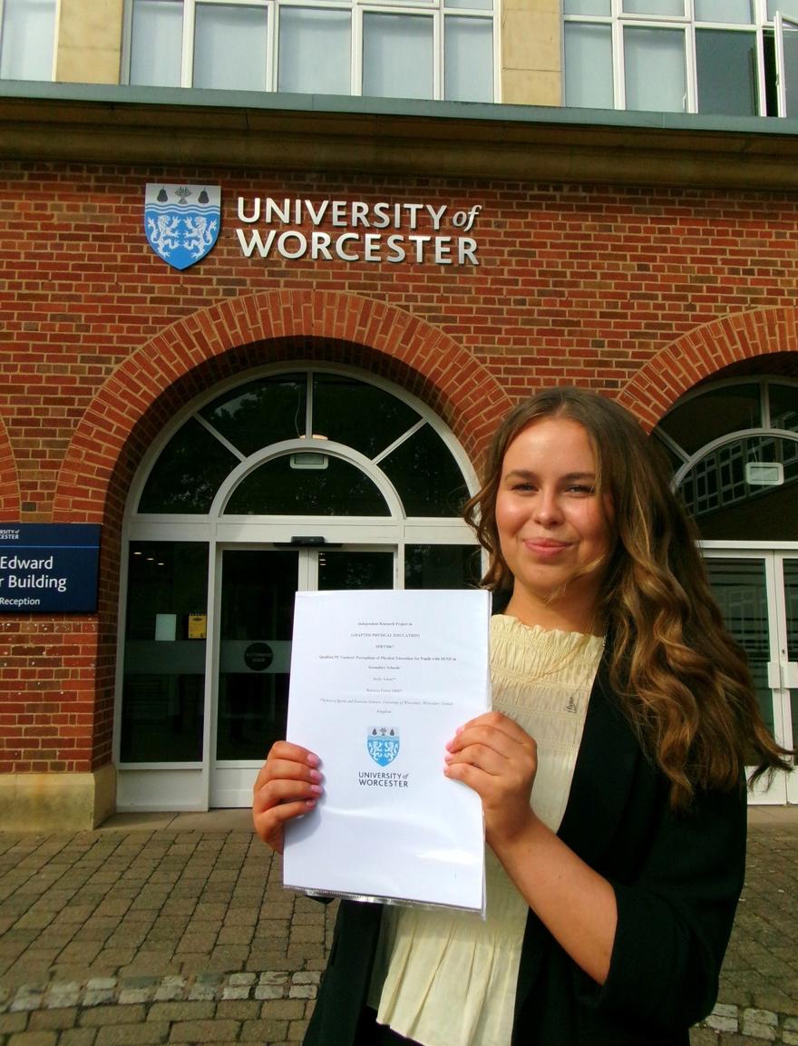 A student poses in front of the University of Worcester while holding a certificate