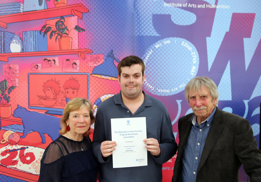 Three people pose in front of a colourful background, the central person holds a certificate