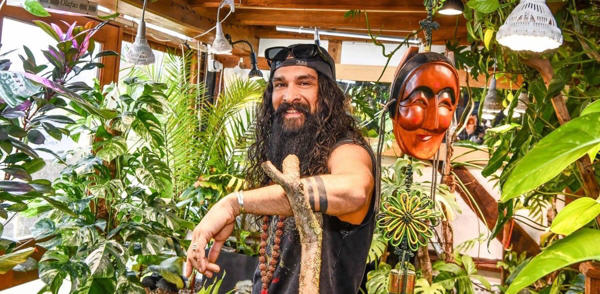 Jonny Balchandani poses amongst plants while leaning on a tree branch, smiling at the camera