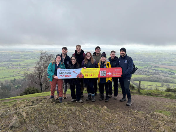 8 Students and a lecturer pose on the Malvern Hills, a beautiful view in the background, as they hold a banner for West Midlands Air Ambulance