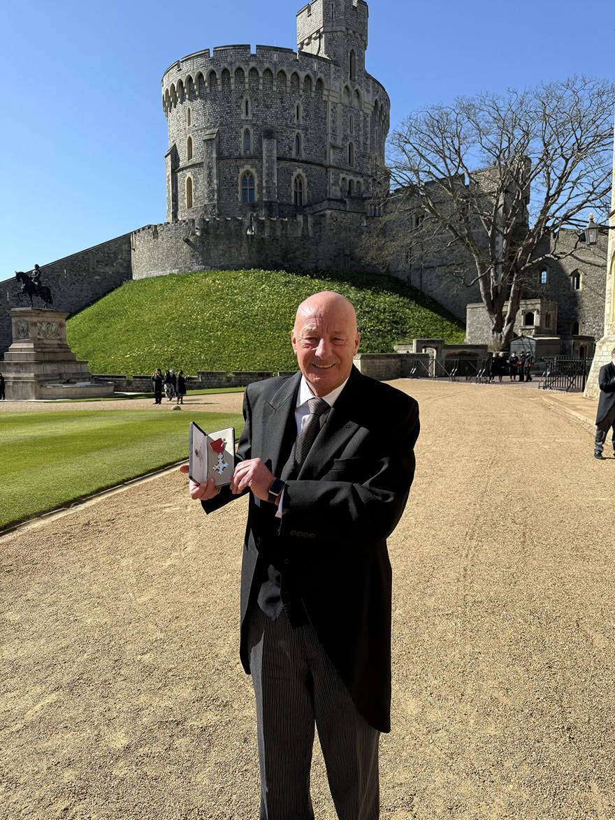 Mick Donovan MBE poses holding his MBE award wearing a black suit in front of Windsor Castle