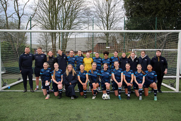 The women's footballers pose as a whole team in a goal with coaching staff
