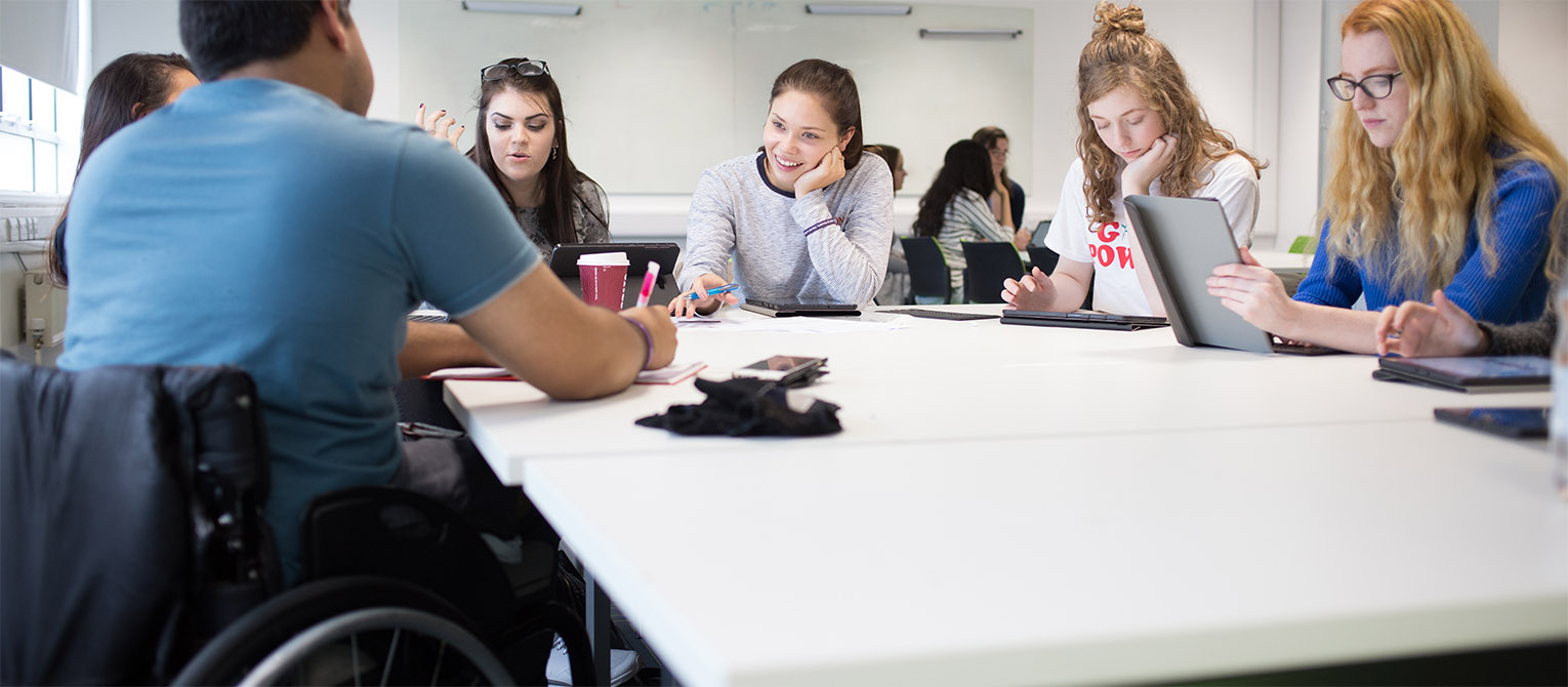 A group of students sat discussing work around a white table in a classroom. One of the students is sat in a wheelchair.