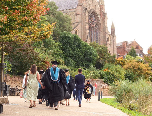 Students in graduation robes walking along tye river with the Cathedral in front of them