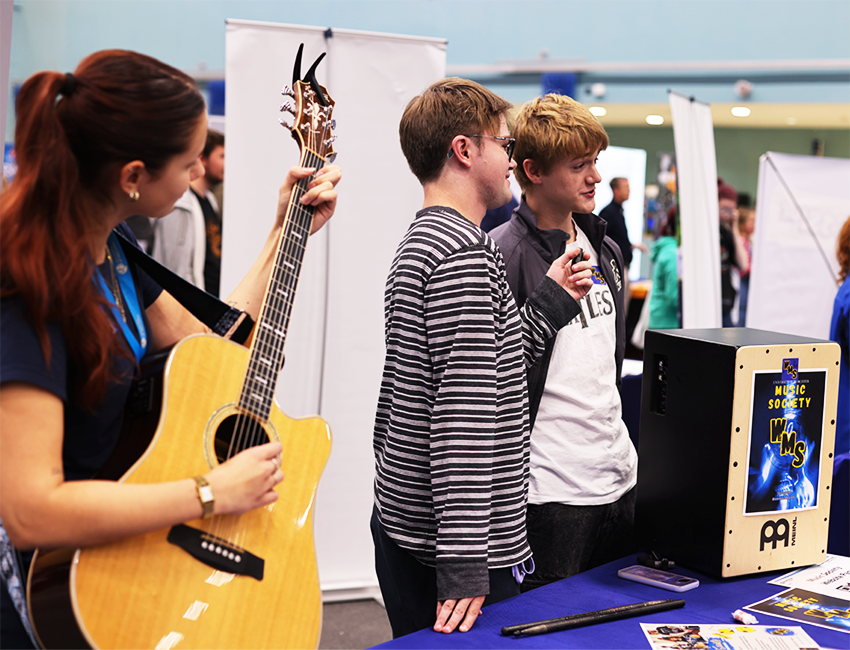 A girl plays guitar whilst two boys speak to students about music society