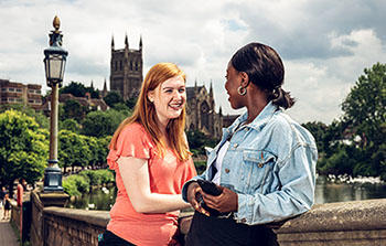 2 students stood smiling and facing each other on Worcester bridge, with Worcester Cathedral in the background