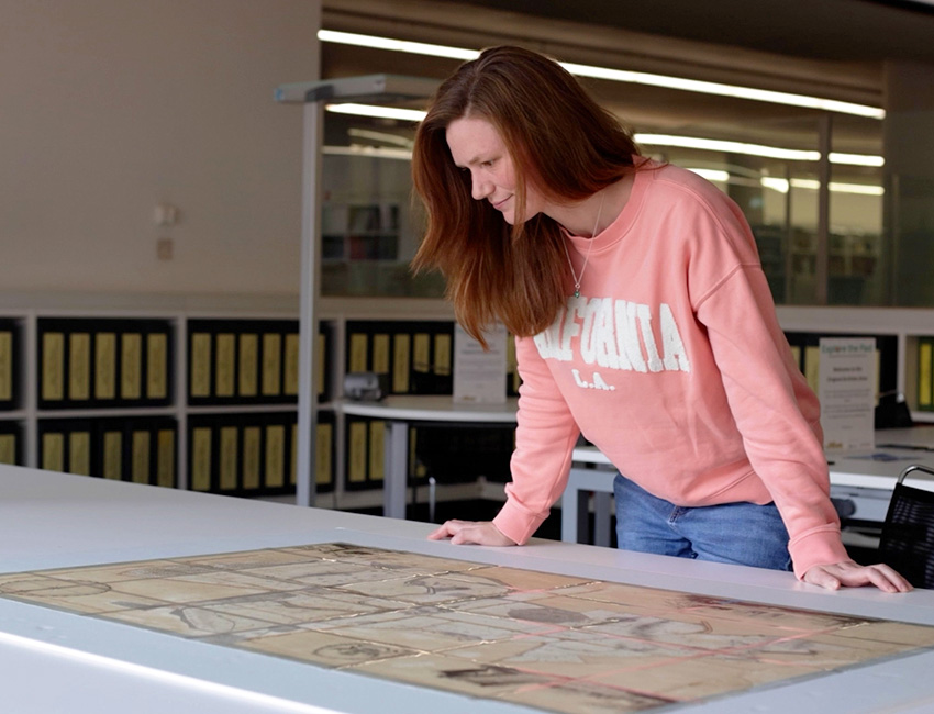 Student examining a historical map in the Hive Library, surrounded by shelves of books and documents.