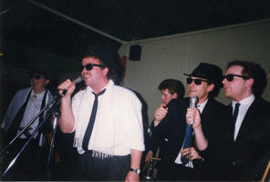 A group of students in sunglasses and hats singing at a party in the 1980s