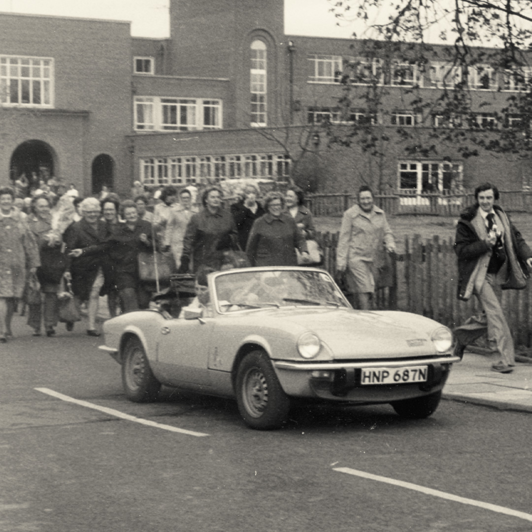 Vintage convertible car driving on a street with a crowd of people walking behind