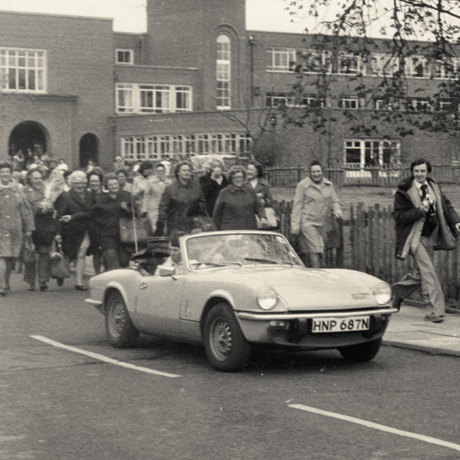Vintage convertible car driving on a street with a crowd of people walking behind