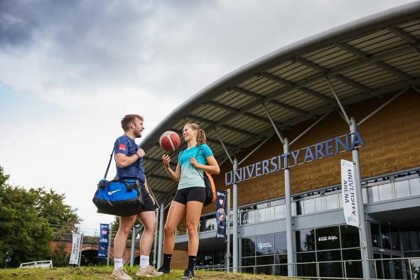 Two students are stood outside the University Arena, dressed in sports attire. One student is holding a basketball.