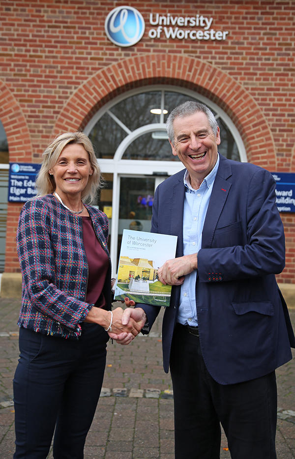 Two people - Dawn Haywood and David Green - standing outside the University of Worcester's main entrance shaking hands and holding a copy of a book about the history of the University