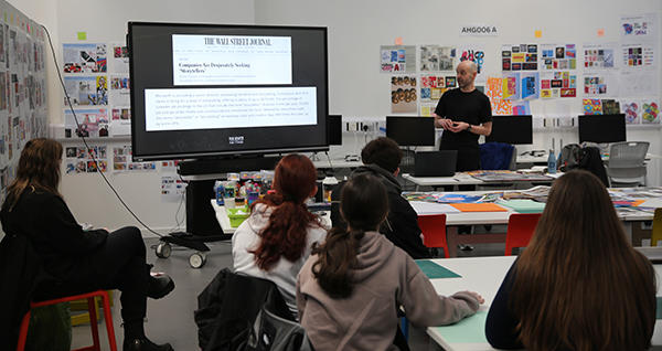 Students sit with their backs to the camera listening to  artist Rob Draper, who is delivering a presentation on a large screen at the front of the room.