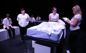 Students acting out a scene in a hospital. One student sits up in a bed while another has a clipboard in hand and two others look on.