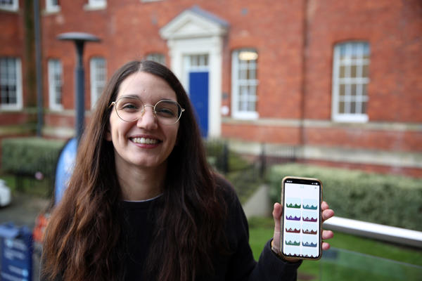 A person holding a mobile phone displaying an app they have developed, standing outside the University of Worcester's City Campus, a large red-brick building