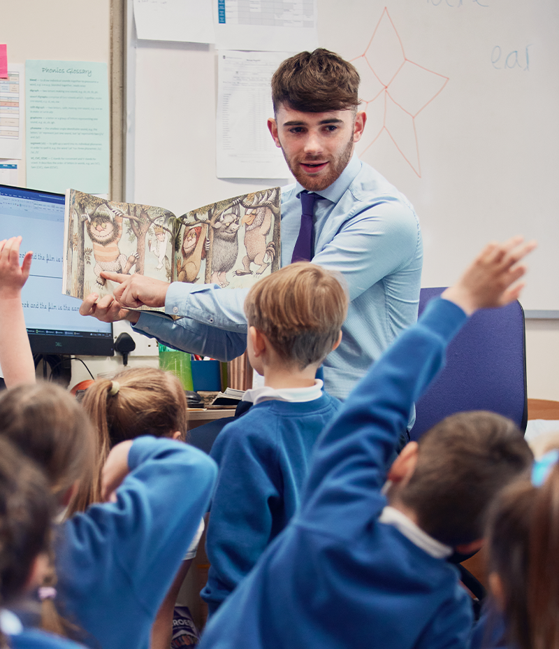 a student teacher holding up an illustrated children's book to a group of young school children