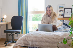 a smiling student is sitting on a bed in University accommodation