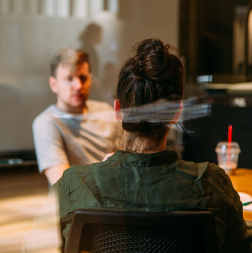 A woman and a man are talking in a coffee shop. She is facing away from the camera and he is facing towards it.