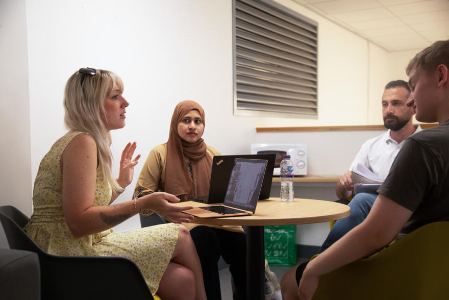 Students sat around a table having a discussion in a social space on campus with microwave in the background