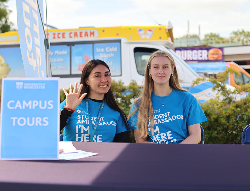 Two students sat behind a table in blue student ambassador t-shirs smiling and waving