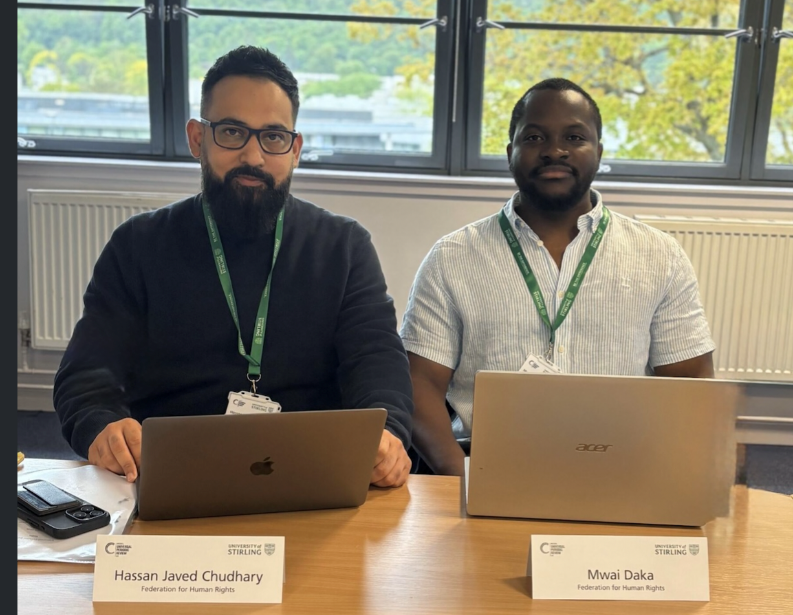 Hassan and Mwai sat at a desk with name cards and wearing green lanyards
