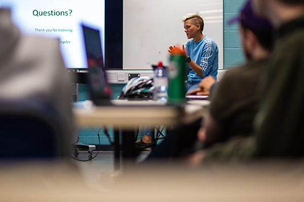 An Adapted Sports lecturer is sitting in front of a class. Behind the lecturer is a screen with text on it.