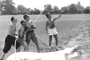 Four shirtless men wearing fireman's helmets and using a hose