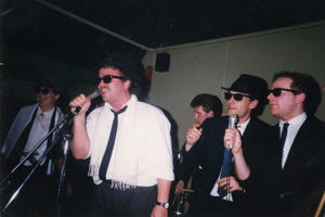 Five men wearing sunglasses and suits are singing into microphones in the 1980s