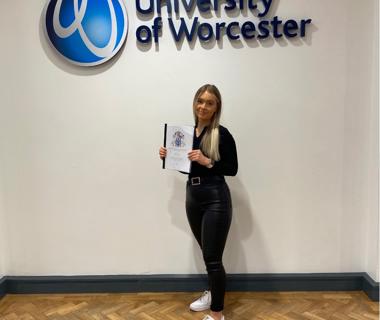 Criminology student stood in front of the University of Worcester logo holding her dissertation project