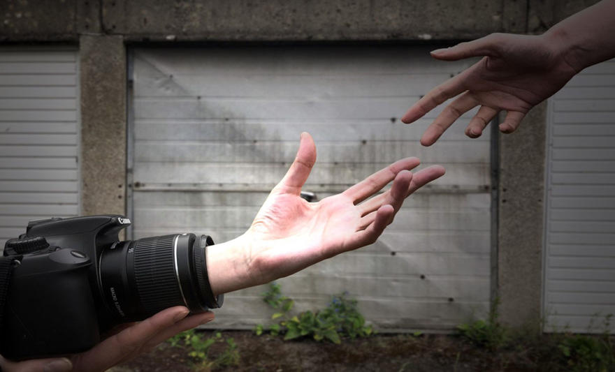 A photo of two hands reaching for each other. One hand is coming out from a camera.