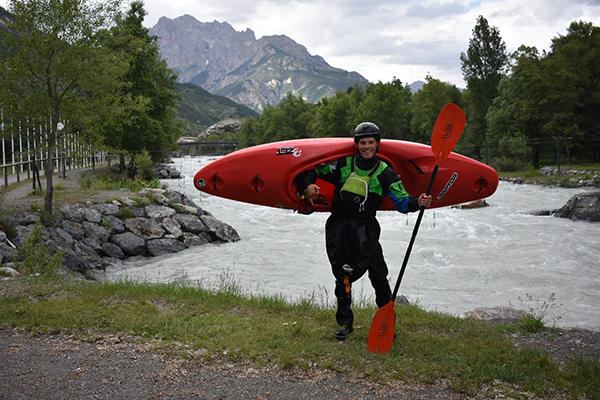 Harry Turner is standing next to a river with choppy waters. On his back he has a red kayak and he is carrying a red paddle.