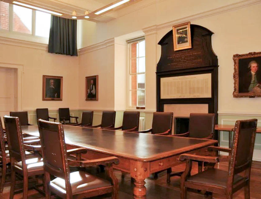 A large wooden table with wide wooden chairs around it, sits in the center of a room with portraits hung from the surrounding walls