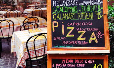 the interior of an Italian restaurant, a sign displaying a menu for different food items is in front of several tables covered in yellow, white and red table cloths.