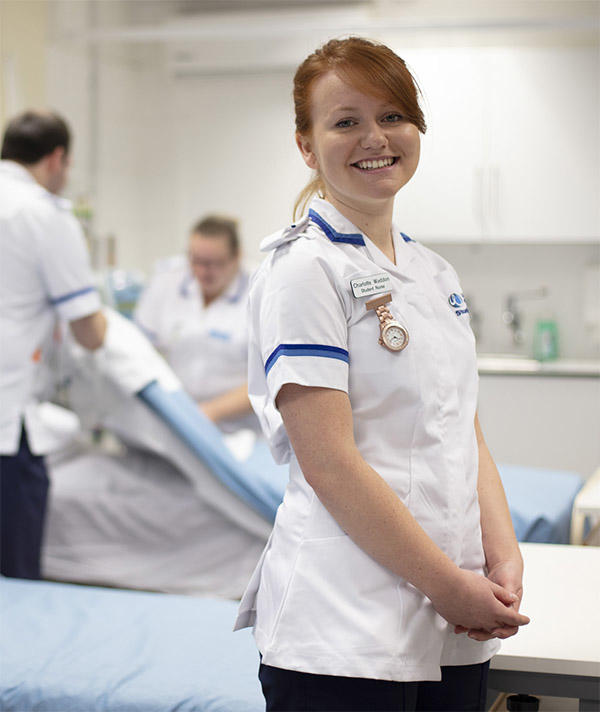 A student nurse smiles while two other nurses make a bed behind her